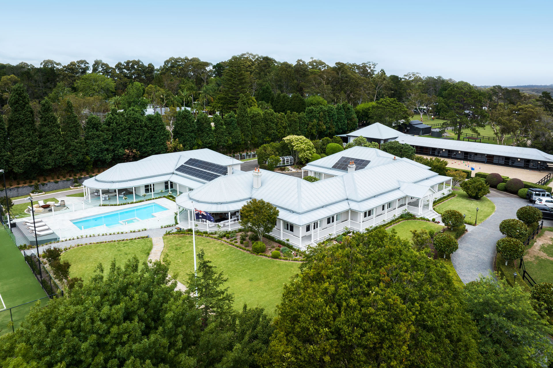 Aerial view of Joalah Ranch, an expansive estate with multiple white-paneled structures featuring gabled roofs and solar panels. The property includes a large swimming pool, meticulously landscaped gardens, a tennis court, and is surrounded by lush, dense greenery. The elegant design of the buildings harmonizes with the natural landscape, embodying a blend of luxury and environmental consciousness.