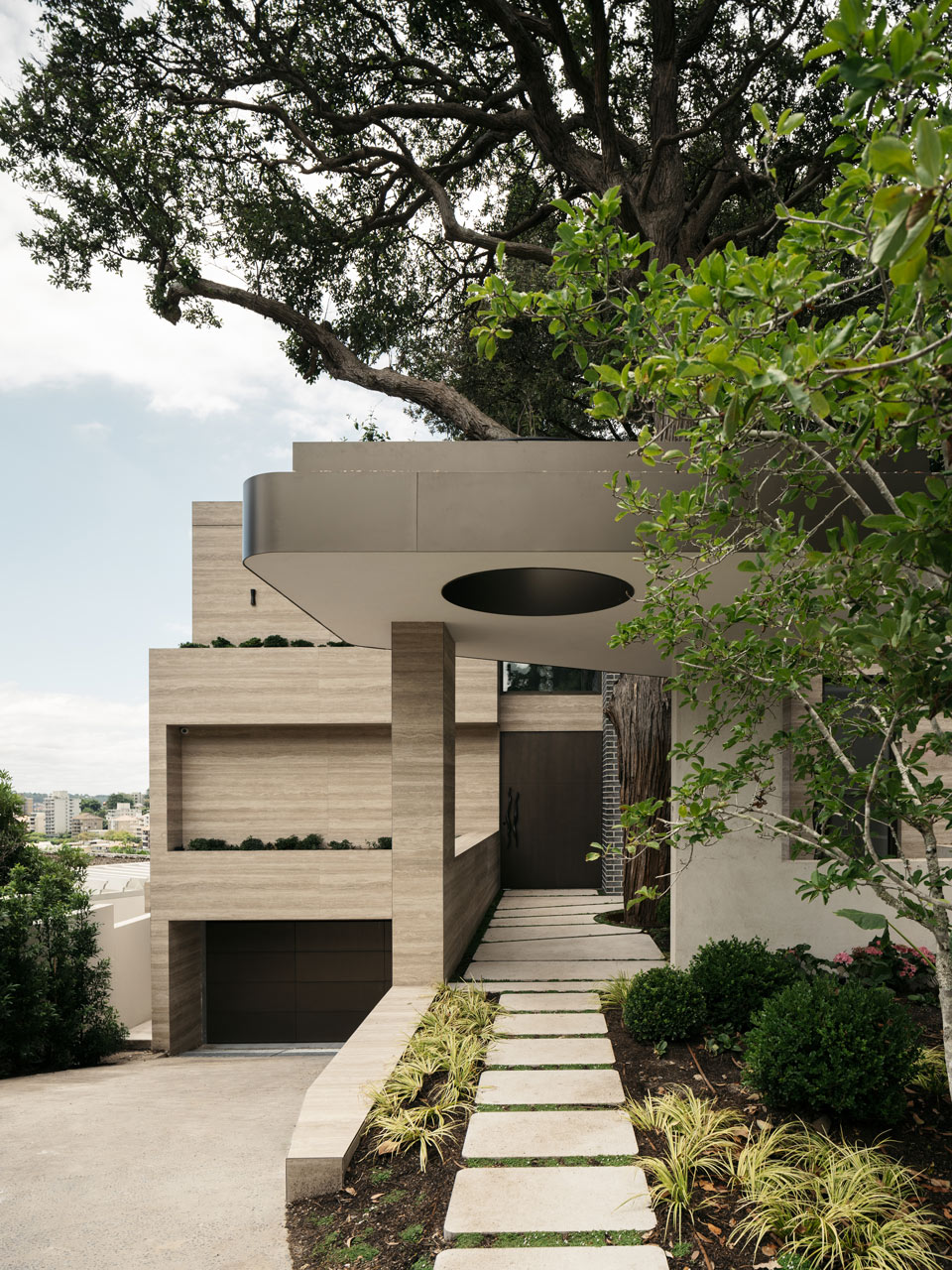 Modern architectural home with textured beige facade, featuring a circular cut-out overhang and a pathway lined with stepping stones and lush greenery.