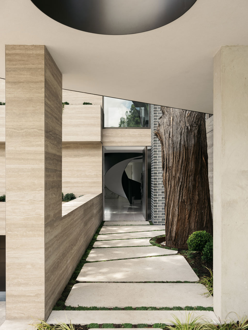 Close-up view of a contemporary building's entryway with a large circular cut-out in the overhang, complemented by a patterned walkway and natural wood textures against a mature tree trunk.