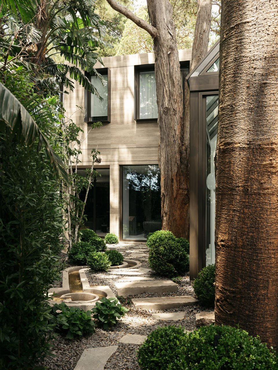 Serene garden pathway leading to a modern house with wood paneling, framed by tall trees and lush foliage, showcasing a harmonious blend of architecture and nature.