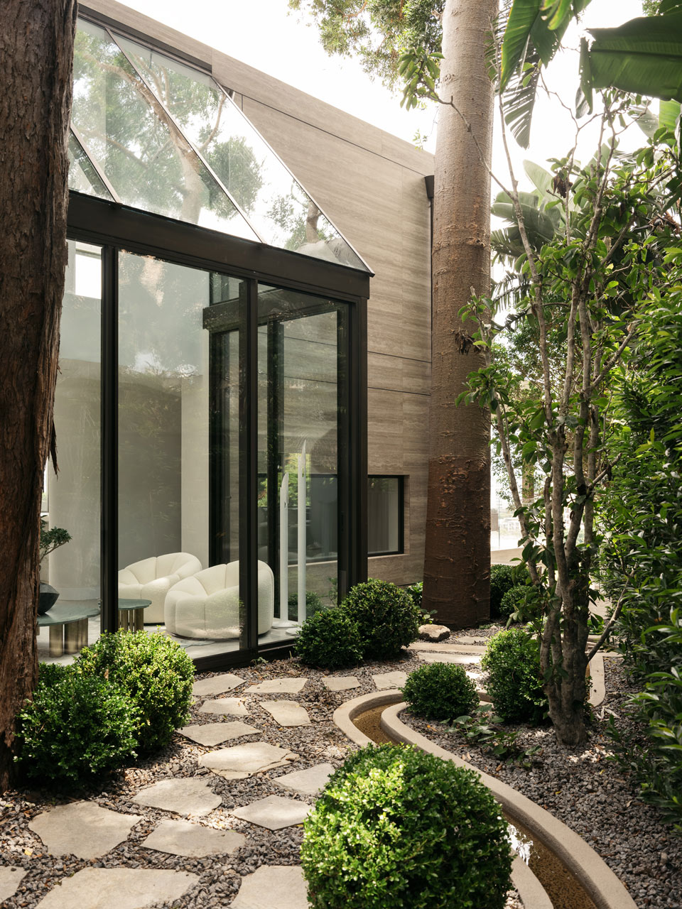 Angled view of a glass corner window of a wooden textured house, integrated with nature featuring a stone pathway and manicured bushes under the shade of tall trees.