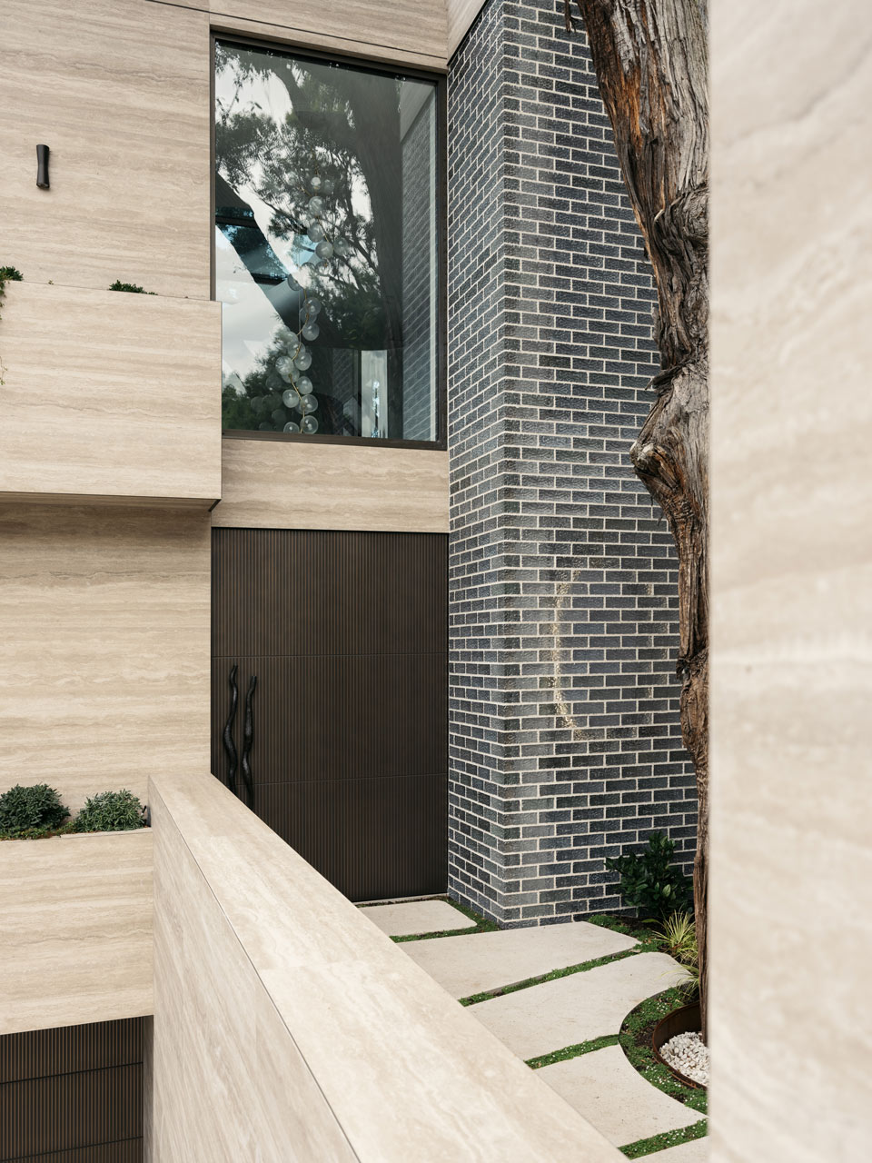 Detail of a building's exterior combining natural wood textures and dark brickwork, with a reflective window capturing the surrounding trees, and a sculptural art piece visible in the niche.