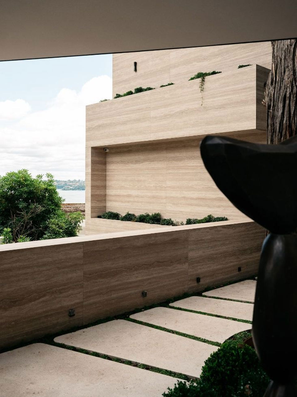 Perspective view from a garden, looking out to a layered wooden façade with integrated planters, under a clear sky, partially framed by a silhouette of a sculpture.